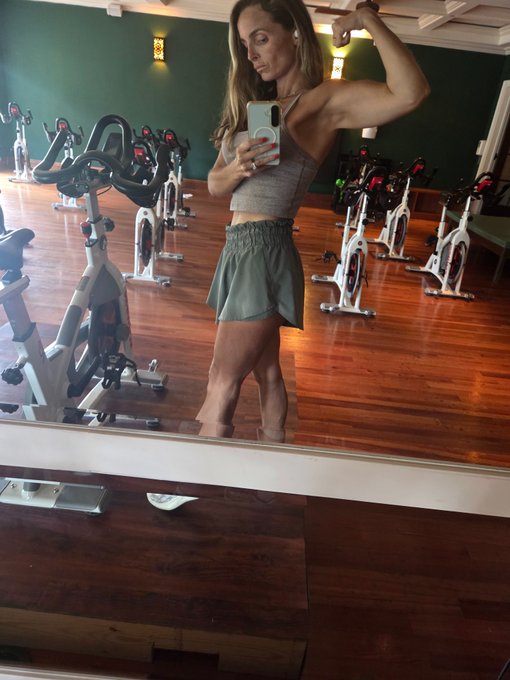 A woman in a gray tank top and green shorts poses for a mirror selfie with a smartphone in what appears to be a spin studio with stationary bikes.