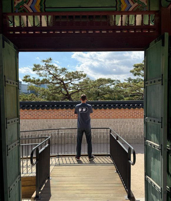 A man in a gray t-shirt stands on a wooden viewing platform framed by ornate traditional Korean gates, looking out over a brick wall toward pine trees and a distant skyline. The setting reflects the "Goodbye, Korea" moment in the post, capturing a final scenic view before heading to Singapore.