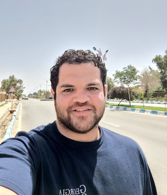 A man with curly hair and a beard takes a selfie outdoors on a sunny day, smiling with a clear road and trees in the background, while wearing a dark shirt with partially visible reversed text reading "GEORGIA," reflecting his morning walk to clear his mind before sharing thoughts about the shipping industry and @EthraShip.
