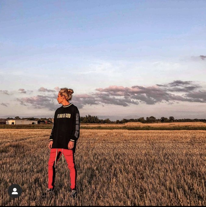 A person with blonde hair tied up stands in a golden wheat field at sunset, wearing a black long-sleeve shirt reading 'FEAR OF GOD' and pink pants.