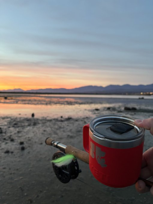 A hand holds a red insulated tumbler and a fishing rod with a green lure, set against a sunset over a calm beach and distant mountains.