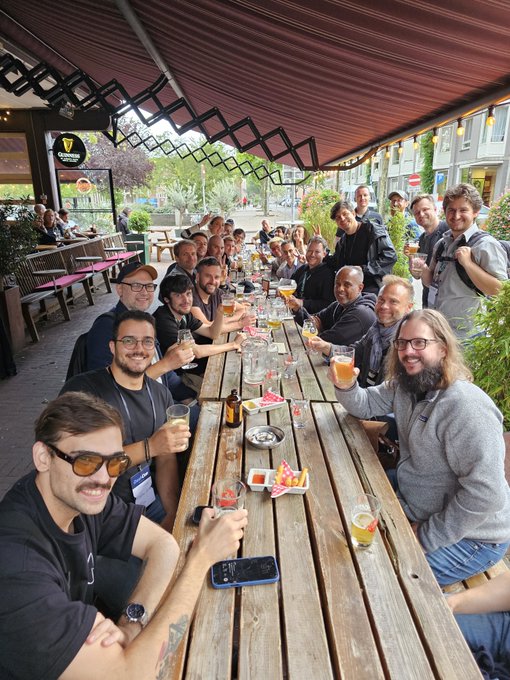 A large group of people sit together at a long wooden outdoor picnic table under a retractable awning, raising glasses of beer for a toast.