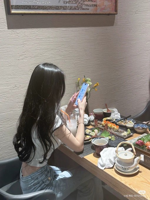 A woman with long black hair sits at a restaurant table filled with sushi and tea, photographing the spread with her smartphone. The image creates an ironic contrast to the post about Bitcoin mining—while the caption discusses miners being "eliminated from the market" during low difficulty periods, the woman is literally observing a meal before enjoying it, mirroring the "eat while you can" sentiment of surviving weak miners before the cycle bottom.