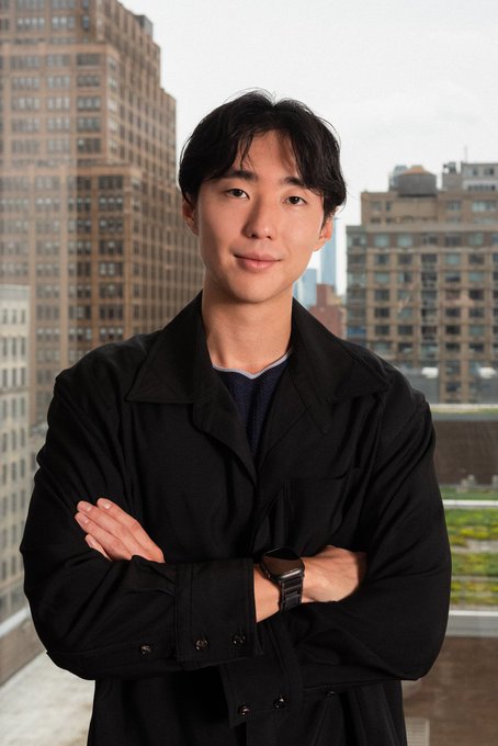 A young man stands on a rooftop with arms crossed, wearing a black coat and smartwatch, with city buildings behind him.