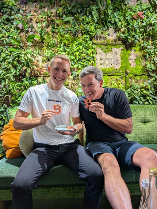 Two men, one wearing a "BITCOINER" t-shirt, smile while holding up Bitcoin logo-shaped cookies and a plate of treats, perfectly illustrating the caption about "two Bitcoiners eating cookies together."