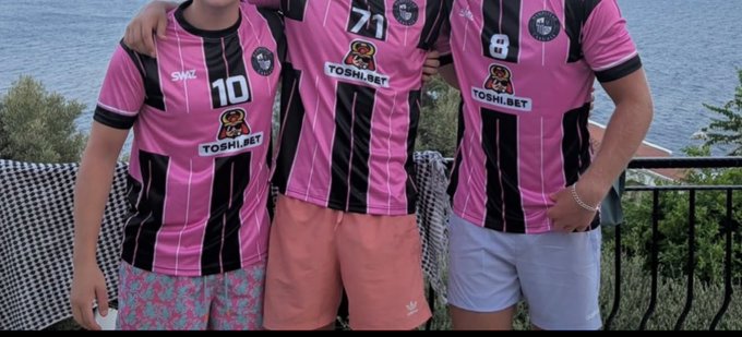 A photograph of three people wearing matching pink and black striped soccer jerseys featuring a dog mascot logo and 'TOSHI.BET' text, standing on a balcony overlooking the sea.