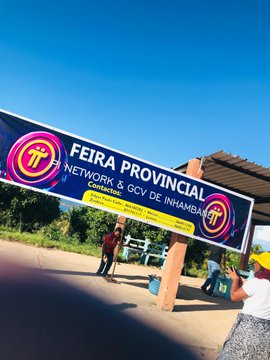 The image features a banner that says "Fiira Province" written in yellow letters against a blue background. The banner also includes a logo for the Pi Network and the GCV Industrial Alliance. In the foreground, there are two people standing next to a bench on a street.