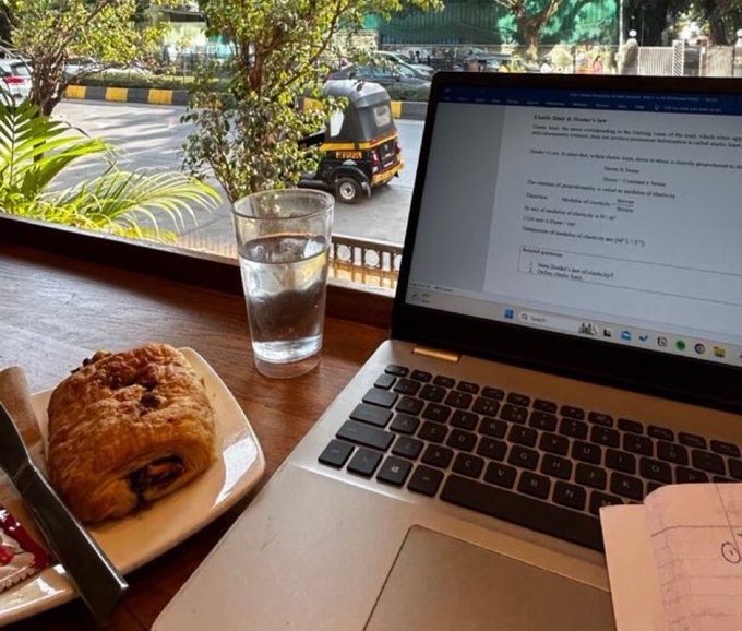 A workspace scene showing a laptop displaying study notes about "Clausius Clapeyron & Clausius Equation" next to a glass of water and a pain au chocolat pastry, with a street view including an auto-rickshaw visible through the window.