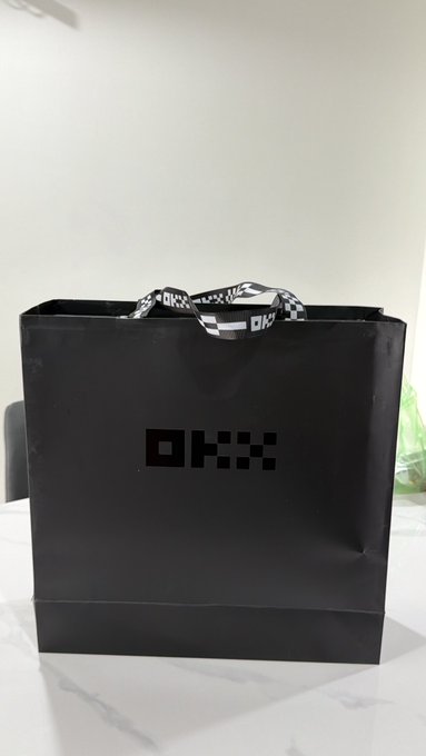 A black paper shopping bag with pixelated 'OKX' branding sits on a white marble countertop.