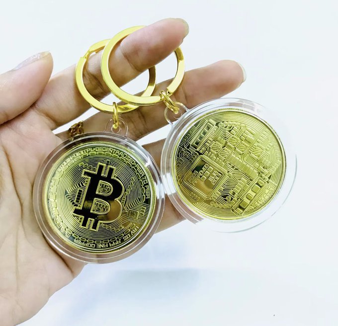 A hand holds two physical Bitcoin keychains, each featuring a gold coin—one showing the Bitcoin logo and the other displaying a circuit board design—encased in clear plastic.