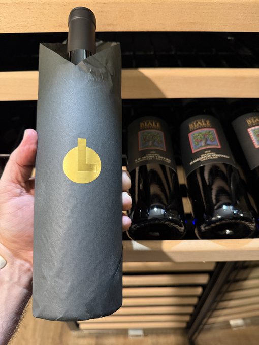 A hand holds a bottle of wine wrapped in dark gray paper with a gold seal, displayed in front of shelves stocked with Riale brand wine bottles.