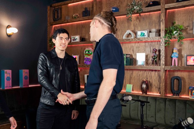 Two men shake hands in a stylish lounge or studio space, with shelves displaying colorful figurines and neon lighting in the background, visually capturing the moment a crypto influencer frames the KOL's bold move to publicly exit his positions and focus entirely on farming Hyperliquid.