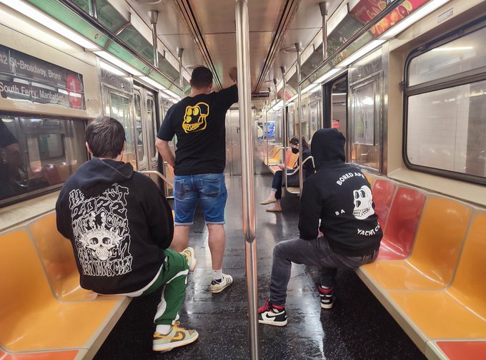 Three young men sit inside a New York City subway car (route 42 to Broadway/Brons/South Ferry), wearing matching Bored Ape Yacht Club hoodies—one with a yellow ape logo and another displaying the distinctive BAYC skull design—capturing the camaraderie celebrated at Apefest 2022.