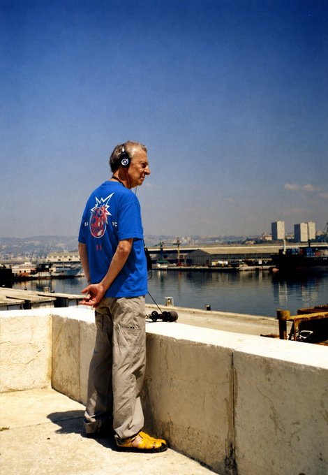 A man in a blue shirt and headphones leans against a concrete wall overlooking a sunny harbor, embodying the post's message about embracing new experiences and hobbies at any age—whether listening to music, exploring a new city, or simply enjoying the moment. The peaceful waterfront scene with boats and distant hills reinforces the idea that it's never too late to find one's own rhythm and join a community of like-minded individuals.