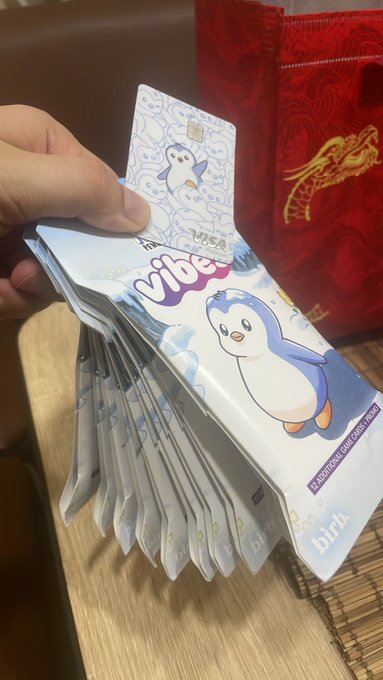 A hand holding a fan of multiple 'vibe' branded Australian debit cards (a 'virgil' card visible on top) featuring a cartoon penguin mascot, displayed against a red fabric background with gold dragon embroidery.