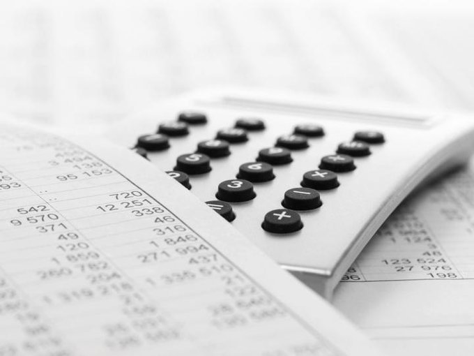 A black and white close-up photograph shows a calculator resting on top of printed financial spreadsheets filled with numerical data.