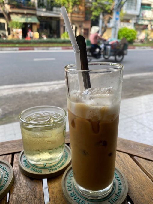 A street-side view of two iced drinks—an iced coffee and an iced lemon tea—on coasters at a sidewalk café in Vietnam.