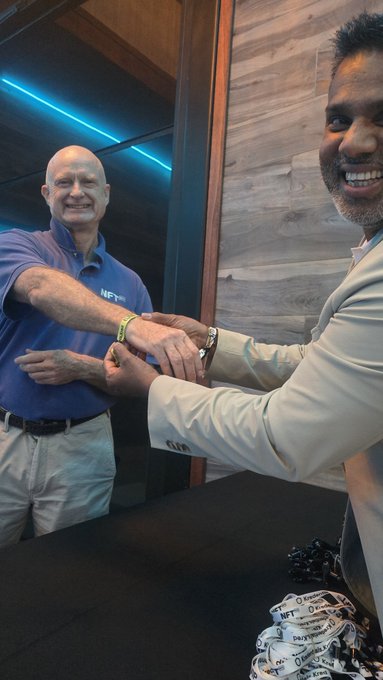 Two men in a wood-paneled room exchange smiles while one places an NFT-branded wristband on the other, with branded lanyards visible on the table.