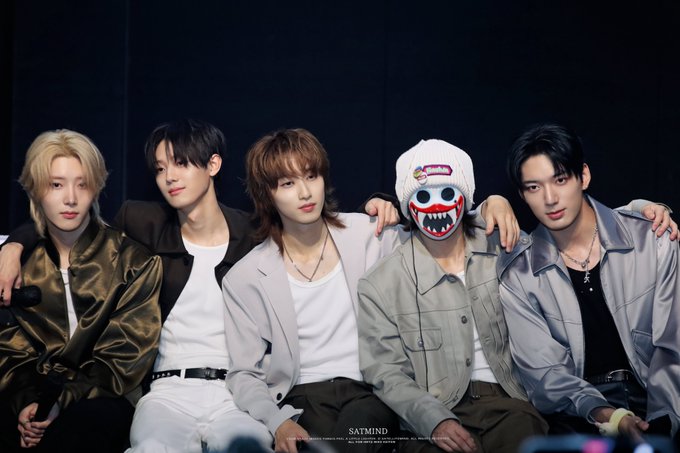 Five young men pose together in stylish outfits against a dark background, with one wearing a white beanie and a shark-mouth mask; the watermark reads 'SATMIND'.