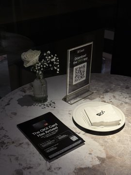 A marble table displays an "OKX Card" invitation alongside a QR code sign prompting guests to download the "OKX" app, a small vase of white flowers, and a stack of napkins on a white plate, capturing the setting of the OKX U Card event.