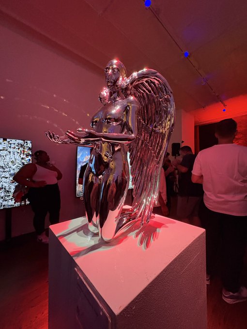 Chrome angel statue displayed on a pedestal in a dimly lit gallery space with pink and blue lighting.