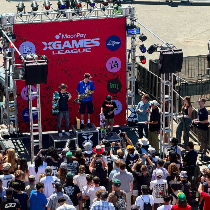 The image shows an awards ceremony at the MoonPay X Games League, where a winner stands on a podium holding up a gold trophy and a skateboard in front of a large red sponsor backdrop. The accompanying caption suggests this is Keith Grossman being credited with handing out the gold medal at the event.