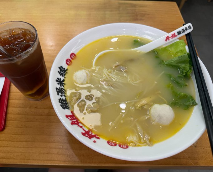 A bowl of soup with noodles, fish balls, and lettuce sits on a wooden table next to a glass of iced tea. The bowl's rim displays Chinese text including '丹旭 鸡油米线' and '酸汤米线', indicating it is a chicken-oil rice noodle dish.