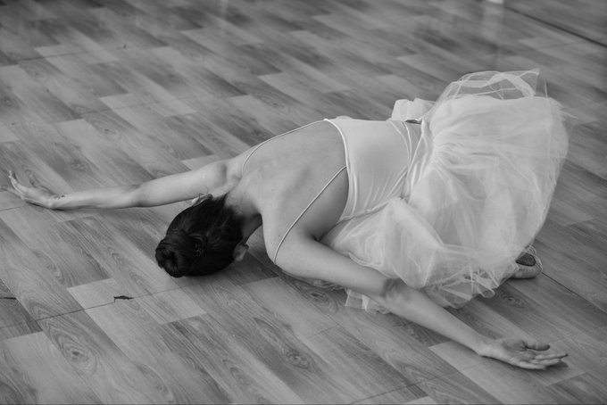 A black-and-white photograph of a ballerina in a white tutu performing an arched pose on a wooden floor, her body curved gracefully with arms extended.