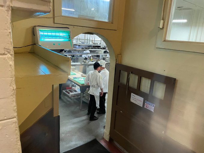 Two workers in white uniforms and hats stand in a commercial kitchen or bakery area with shelving, visible through an arched doorway, with a blue light fixture mounted on the wall.