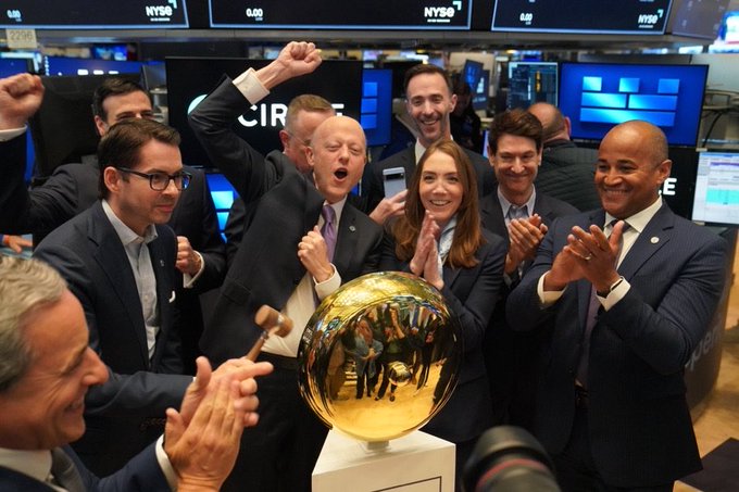A group of people in business attire celebrate enthusiastically at the NYSE, with one man ringing a ceremonial golden bell.