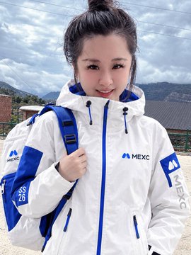 A smiling woman models a white and blue "MEXC" branded jacket with the hashtag "#MEXC26" visible on the sleeve, while carrying a matching backpack outdoors. The image relates to the post as a promotional example of the fashion challenge, showing participants wearing the AI-outfitted MEXC clothing that fans can virtually try on for a chance to win prizes.