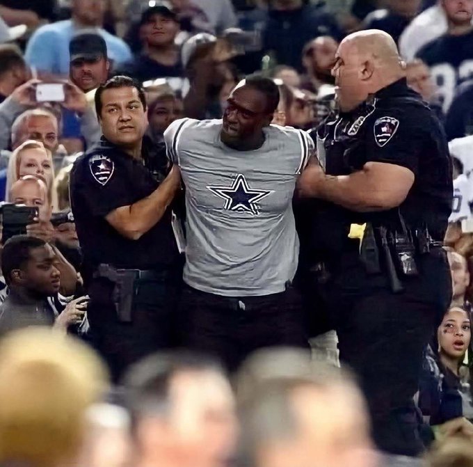 Two police officers escorting a man wearing a Dallas Cowboys jersey through a crowd at a sporting event.