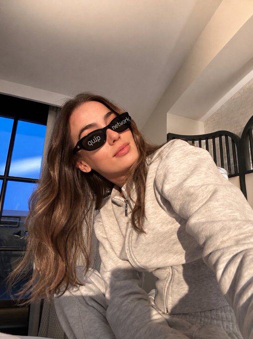 The image features a woman wearing sunglasses, sitting down with her arms crossed over her chest. She has long hair and is dressed in a grey sweatshirt or hoodie. The photo captures a moment where she appears to be posing for a picture while wearing these sunglasses. The caption of the image reads "hii my fam yesterday i talked about the rewards but i didn't explain what @quipnetwork is in the simplest terms: if quantum computers become a real threat one day how do we protect existing wallets and assets? that'S the problem quip is trying to solve their idea is". The woman's pose, along with her sunglasses, suggests she might be participating in an event or gathering related to technology or science.