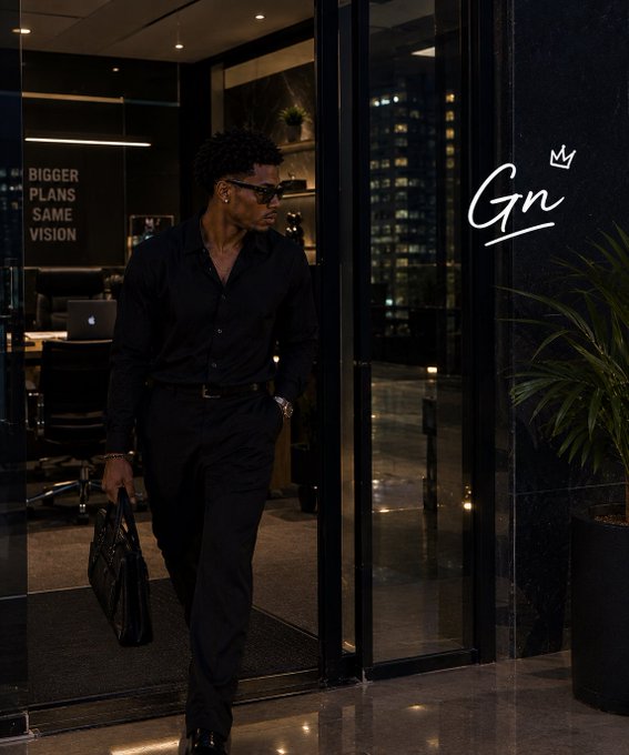 A stylish man wearing an all-black outfit and sunglasses walks through a glass door holding a briefcase, with the text 'BIGGER PLANS SAME VISION' on the left and a 'Gn' logo with a crown on the right.