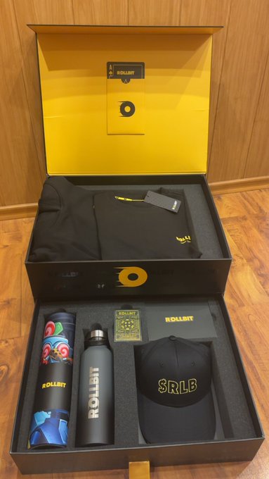 An open Rollbit branded box containing merchandise including a black hoodie, a colorful water bottle, a black cap with '$RLB', and other promotional items.