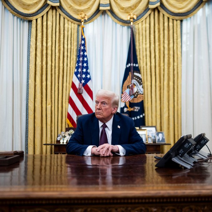 Donald Trump sits at the Resolute Desk in the Oval Office, behind him are the American and Presidential flags. This image is used to humorously represent how influential public figures, rather than market metrics, can impact cryptocurrency trends.
