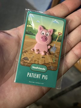 A hand holds a small trading card pack labeled 'Veefriends' and 'PATIENT PIG', featuring an illustration of a pink pig holding a shovel in a field.