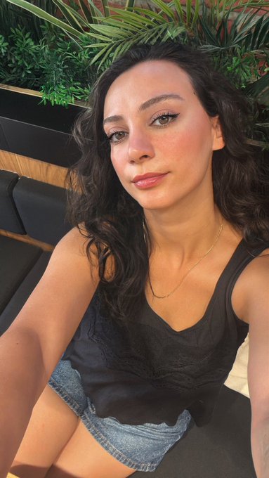 A selfie of a young woman with dark curly hair wearing a black tank top and denim shorts, sitting outdoors with green plants visible in the background.