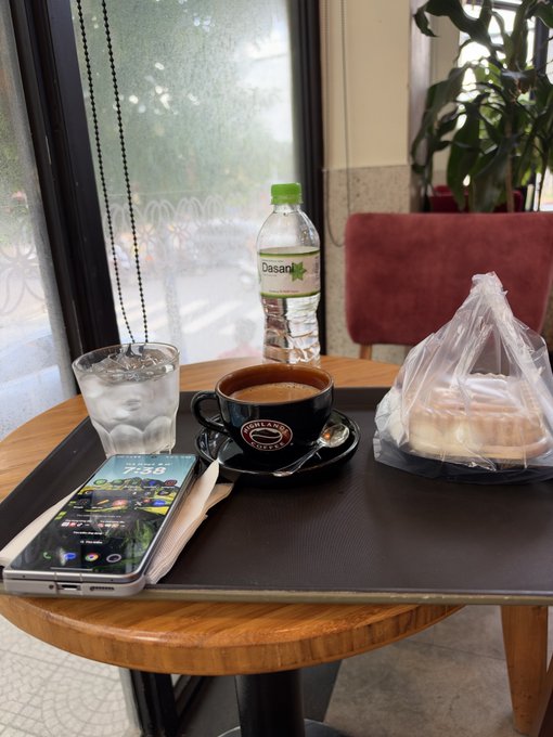 A café table setup featuring a glass of iced water, a bottle of Dasani water, a cup of coffee on a saucer, a pastry in a plastic bag, and a smartphone displaying an app interface.