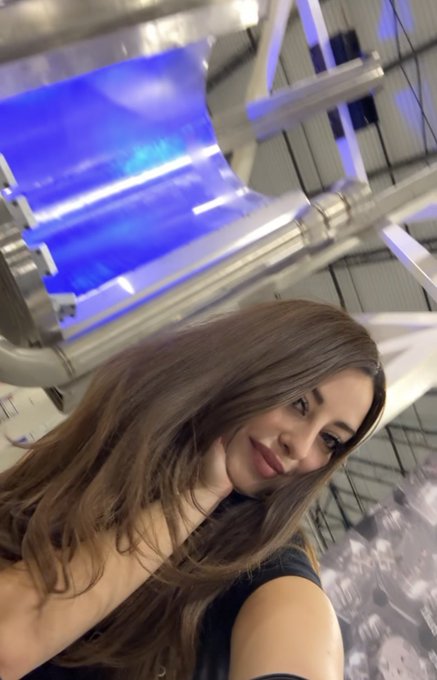 A young woman with long brown hair smiles for a selfie in front of a large industrial nuclear reactor, its glowing blue core visible behind her. The post's caption plays on the scientific setting, suggesting a playful, bold connection between the subject and the powerful machinery looming behind her.