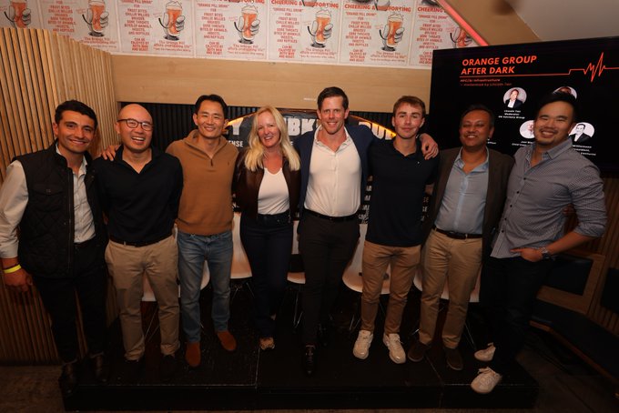 The Orange Group team poses together at their "OG After Dark" event in NYC, standing in front of a branded backdrop displaying the event's logo and tagline "#REWRITINGTHENARRATIVE," with speakers and partners gathered to discuss the convergence of AI, digital assets, energy, and compute.