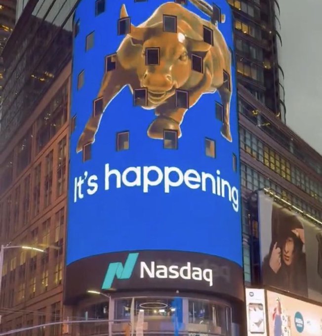 A photo of the Nasdaq building display in Times Square showing a golden bull breaking through a blue background with the text 'It's happening' and the Nasdaq logo.
