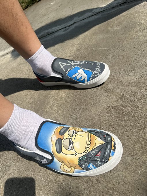 A close-up photo of someone's feet wearing custom slip-on shoes decorated with Homer Simpson graphics and graffiti-style art (including 'AK' and 'HELP'), paired with white socks, standing on a concrete sidewalk.