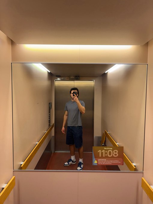A man in a gray t-shirt and denim shorts takes a mirror selfie in an elevator at 11:08 AM on Thursday, August 21, as noted on the digital display, signaling he's stepping away for the day as mentioned in his "Gthursday" post.