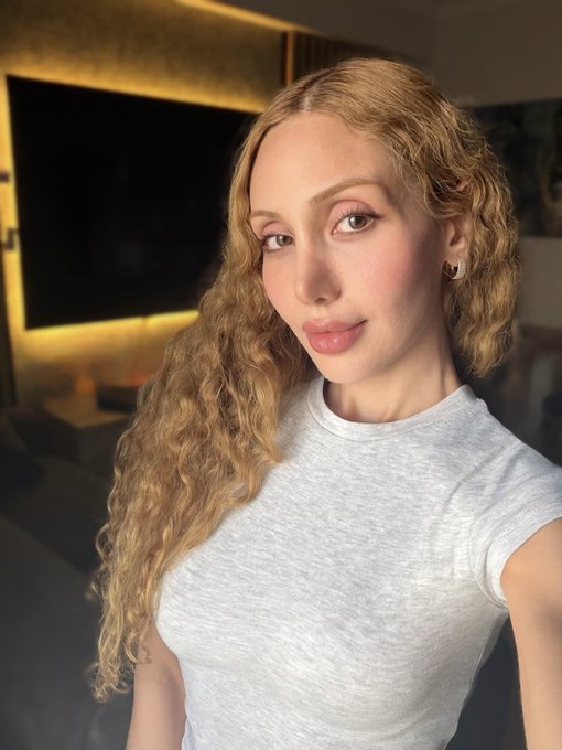 A selfie of a young woman with long, wavy blonde hair wearing a white t-shirt and small hoop earrings, standing in a modern room with a dark TV screen in the background.