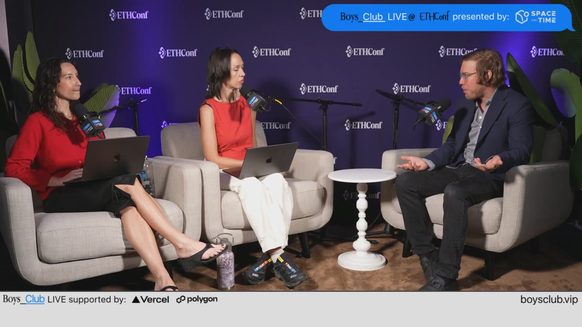 The image shows a panel discussion with three people sitting around a table, each holding a laptop computer. The panelists are engaged in conversation, possibly discussing a topic related to their work or personal interests. A microphone is also present at the front of the room, indicating that they might be recording the discussion for later use. 

The background features a purple wall with logos and text, which could be part of the event's branding or sponsorship. The image also includes some text elements such as "Boys Club LIVE @ ethcon", "@spaceandtime", "#serotoninhq", "@amandacassatt", "@aseemk", and "@chriseyin".