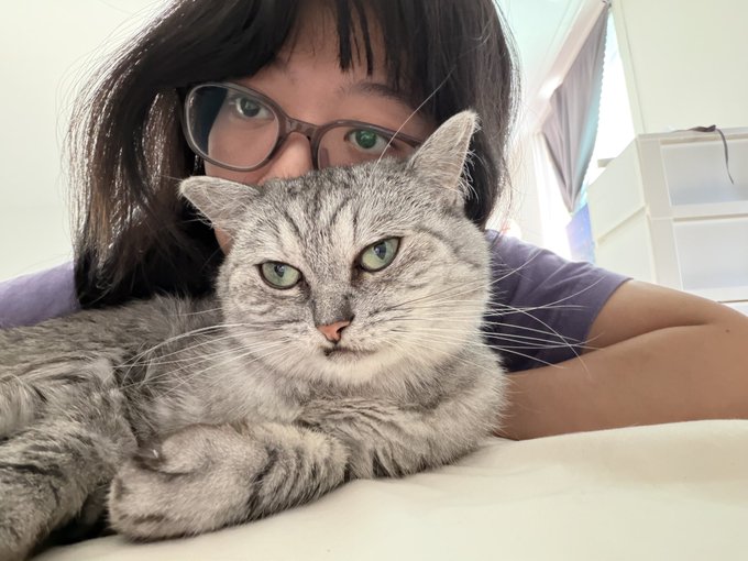 A woman with dark hair and glasses poses closely with her gray tabby cat, both looking directly at the camera.