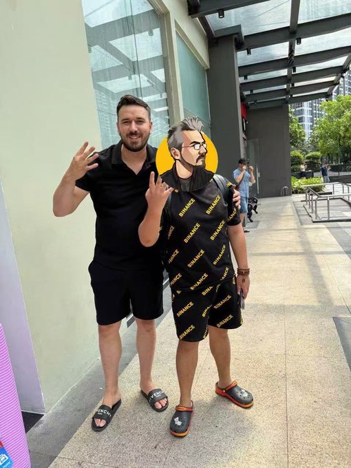 The image features two men standing next to each other, with one of them wearing a black shirt and shorts while the other has a yellow face mask on his head. They are posing for a picture together, possibly at an event or gathering. The background includes a building and some trees, suggesting that they might be in an urban setting. There is also a pink sign visible in the image, which could potentially be related to the event they are attending or simply part of the overall decor.