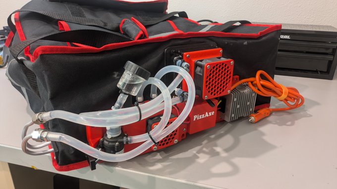 The image shows a custom-built "PizzAxe" device, featuring a Bitcoin mining rig (with the "PizzAxe" name visible on the red casing) connected to clear tubing and heating elements, all packed in a black and red insulated delivery bag used to keep pizzas warm. This invention cleverly combines a pizza delivery driver's two interests—mining $BTC and delivering hot pizzas—by using the miner's heat output to warm food during transport.