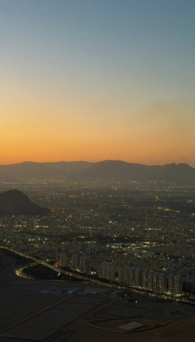 An elevated evening view captures a sprawling city with twinkling lights and highways, set against a gradient orange and blue sunset sky with distant mountain silhouettes.